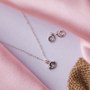 A silver pendant set with a snail motif design, consisting of a pendant necklace and a pair of earrings, displayed on a pink fabric surface.